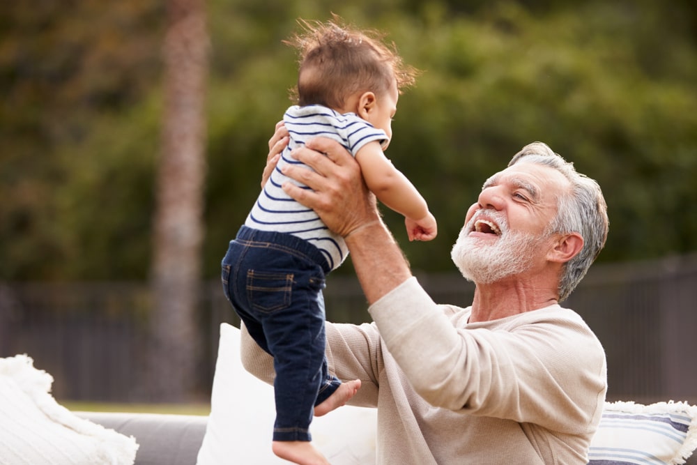 Senior Hispanic man sitting in the garden lifting his baby grandson