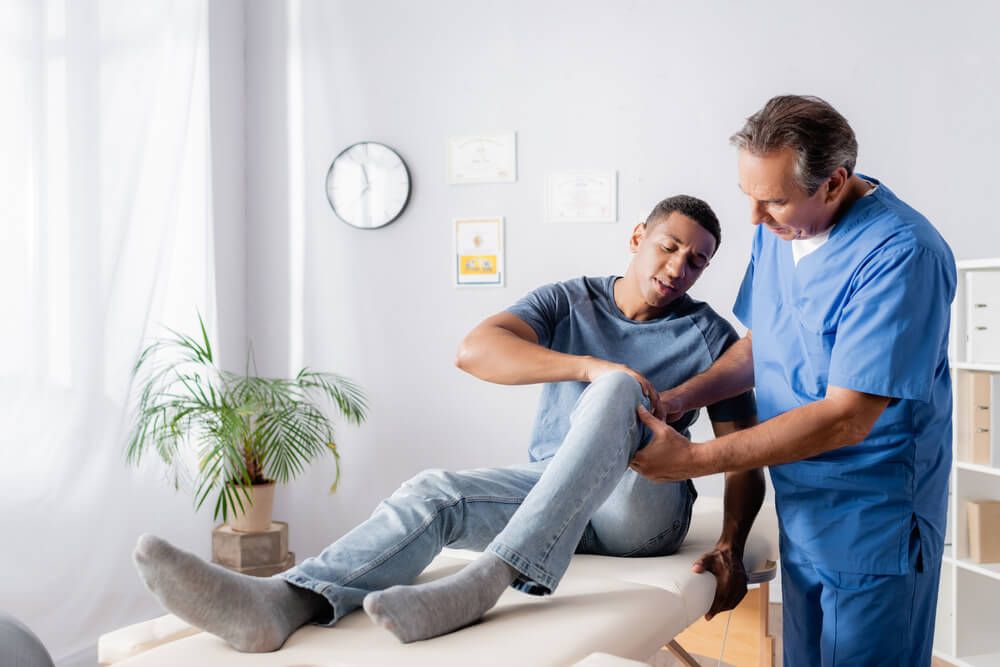mature chiropractor touching injured knee of african american patient