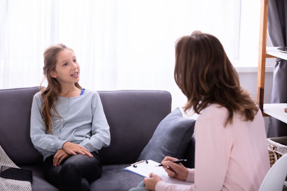 Girl Sitting On Sofa Explaining Her Problems To Female Psychiatrist At Home