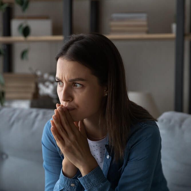 Worried young woman sit on sofa look aside lean forward with folded hands