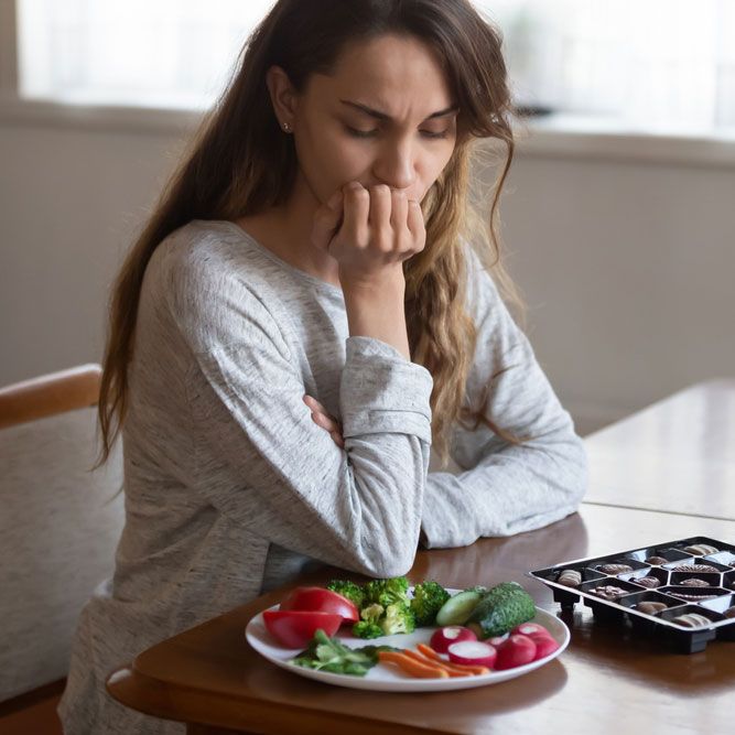 Unhappy young Latin woman look at chocolates and vegetables face temptation suffer from eating disorder.