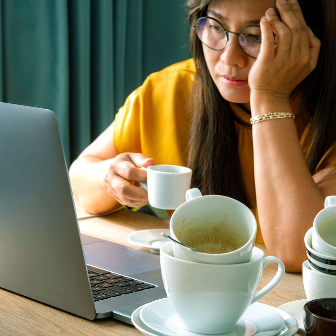 A piles of used coffee cups on desk front of an overtired Asian woman holding a cup of coffee sitting working frustrated exhausted looking at laptop screen.