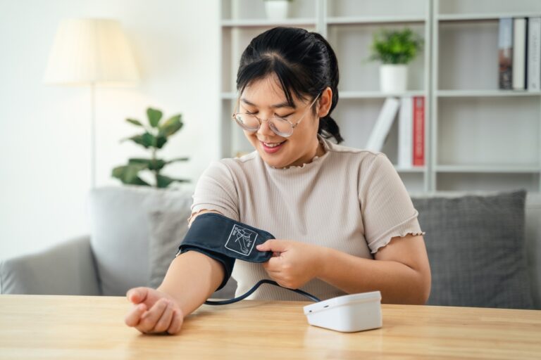 An Asian woman uses a portable blood pressure monitor in her living room. Health awareness concept.