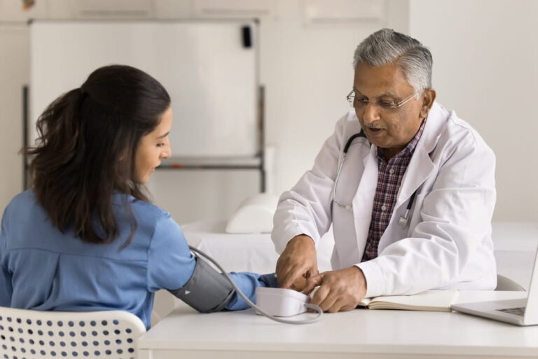 Senior Indian doctor man checking blood pressure of young Latin woman, using digital electronic device, medical instrument, measurement tool. Patient visiting practitioner for examination, checkup