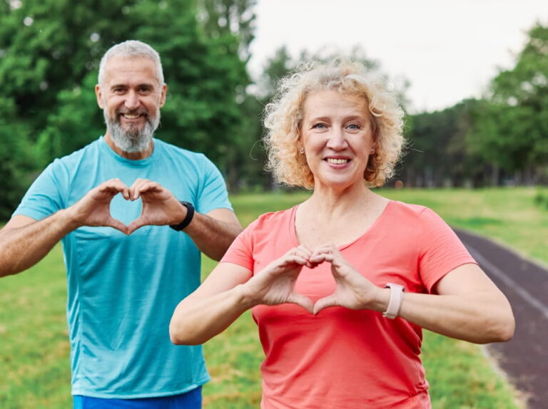 Smiling active mature middle aged couple people posing together forming heart shape with hands in the park