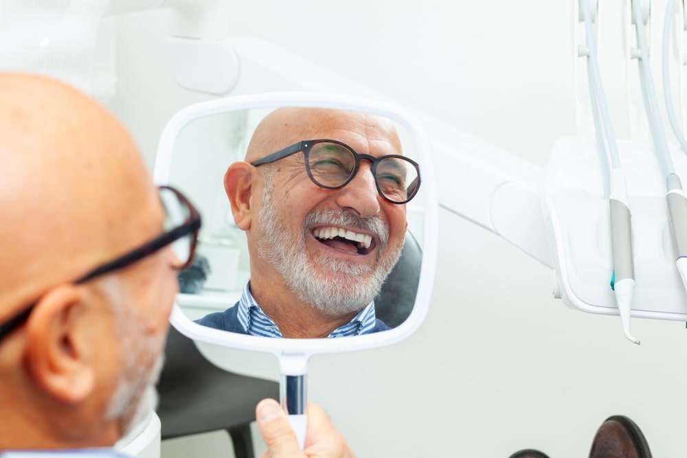 Happy senior patient admiring his perfect smile after a dental implant treatment, holding a mirror in his hand at the dental clinic