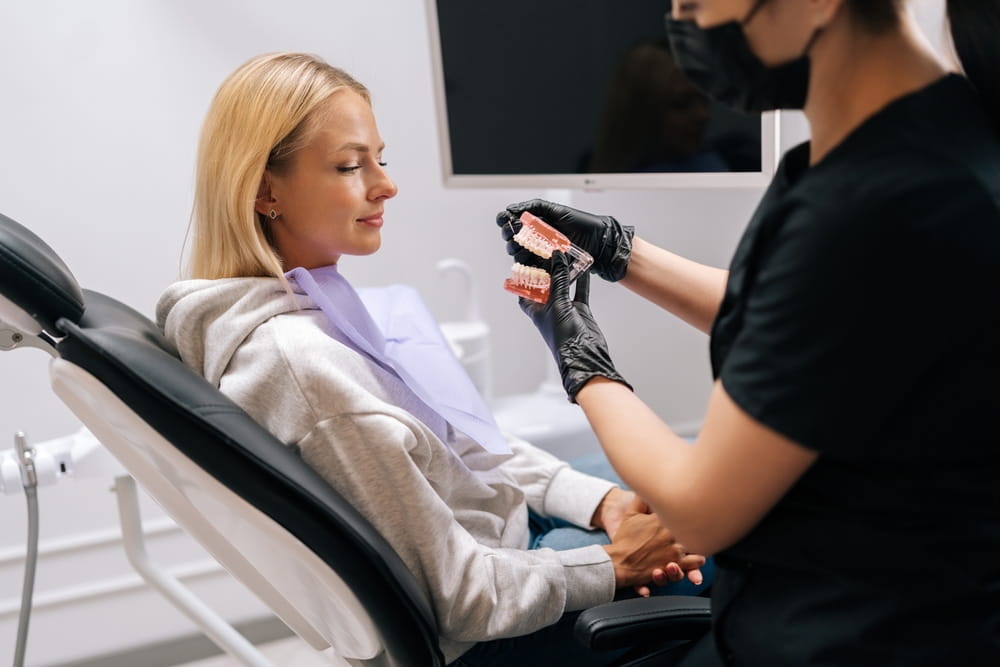 Side view of unrecognizable female dentist dentist in protective mask and gloves showing jaw model with braces to young woman patient in dental office