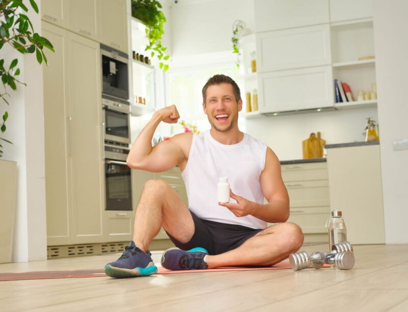 A fit and muscular man displays his strength by flexing his bicep and holding a jar of vitamins, tablets, or dietary supplements in the other hand, while seated on a workout mat at home.