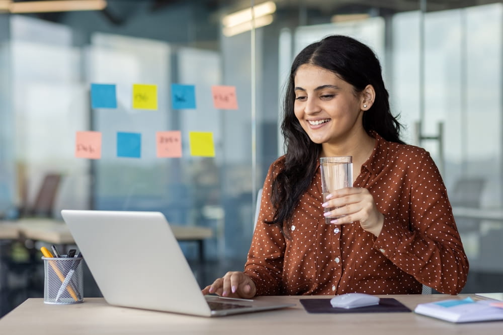businesswoman holds a glass of water, smiling at laptop in office. Image highlights hydration, focus, and positivity in work