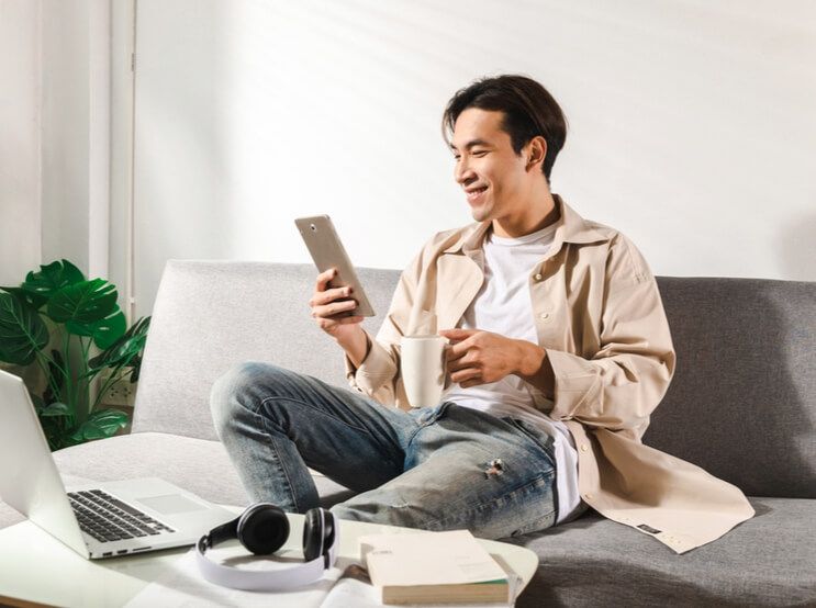 Happy smiling Asian student sits and relaxes on a couch using modern tablet