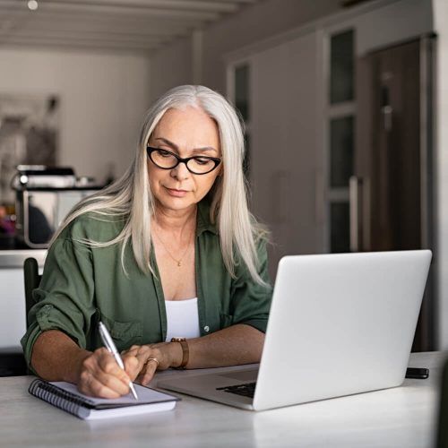 Senior stylish woman taking notes in notebook while using laptop at home