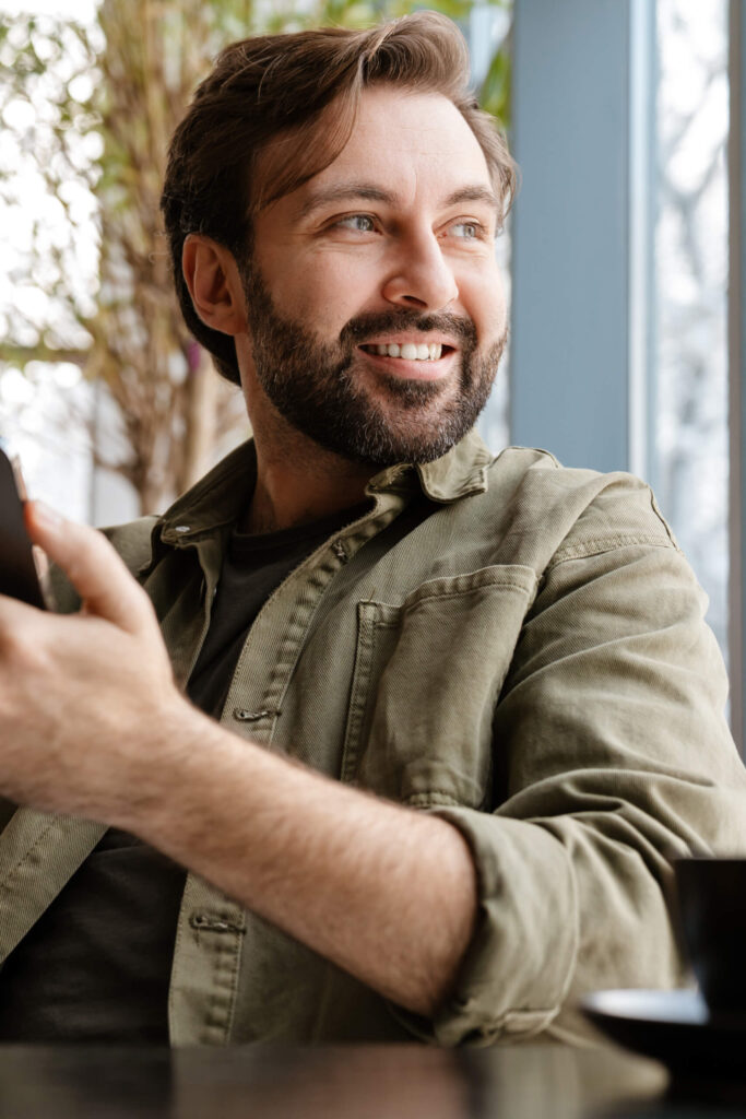 Unshaven happy man smiling and using mobile phone while sitting in cafe