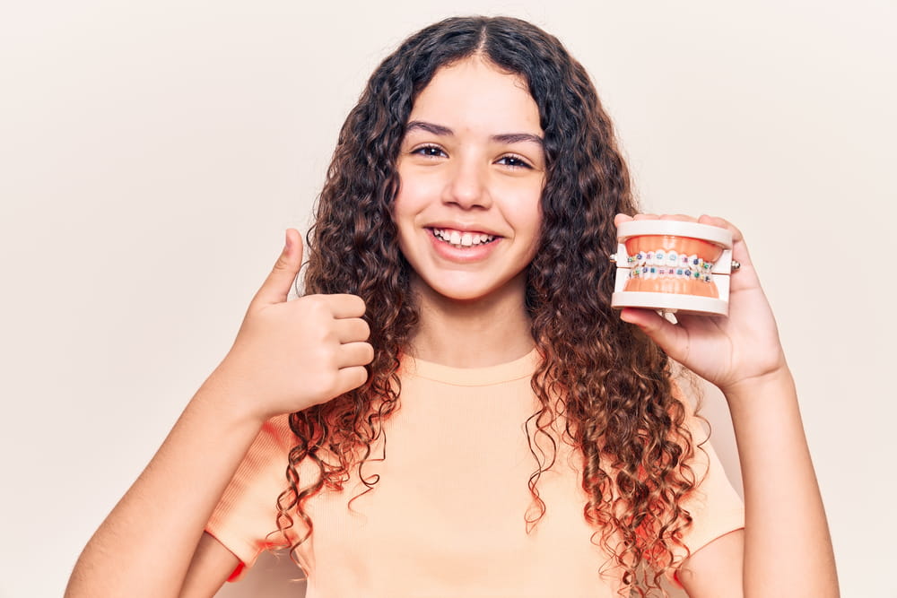 Beautiful kid girl with curly hair holding orthodontic smiling happy and positive, thumb up doing excellent and approval sign