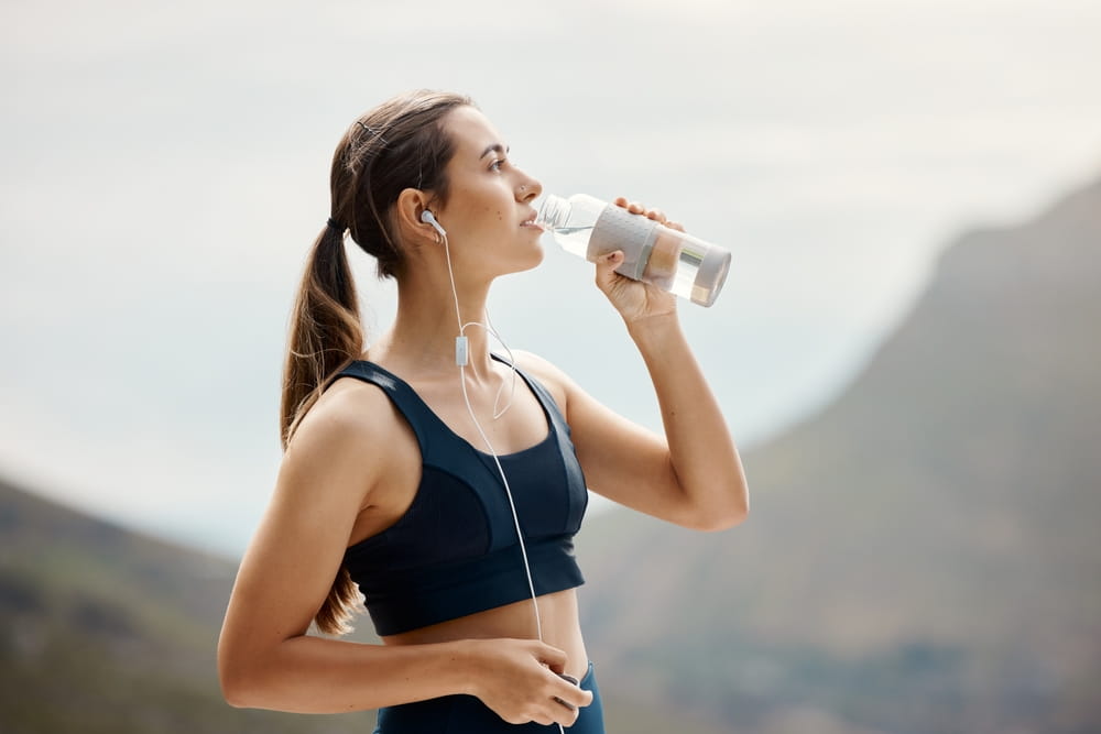 Fitness, woman with earphones, drinking water and hiking in nature for health and wellness during exercise