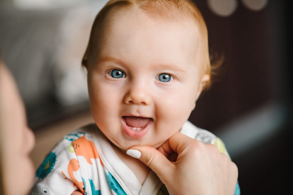 Baby boy portrait looking at camera. Close-up mother hands open baby mouth to examine first teeth. Infant primary tooth.