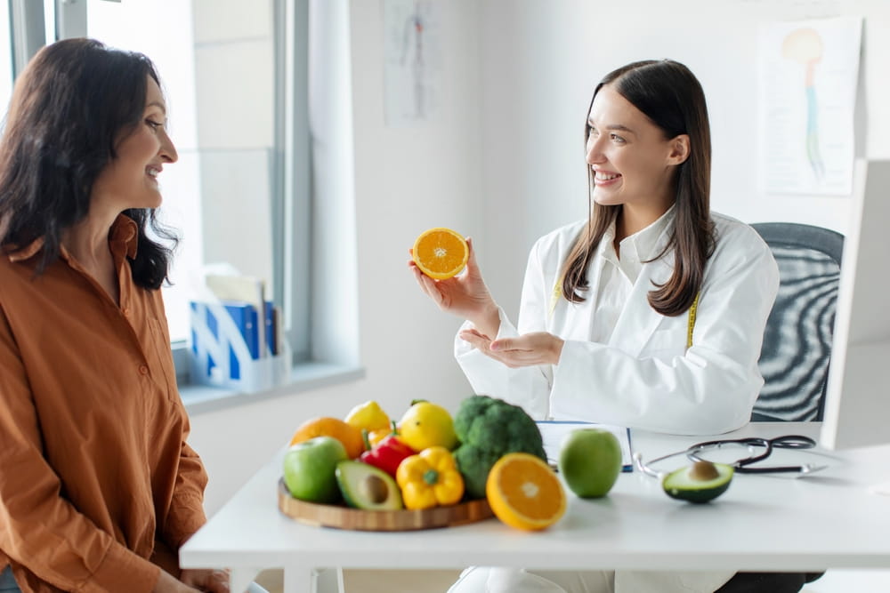 Nutritionist woman giving consultation to mature female patient, holding half of orange and talking about nutrition and diet